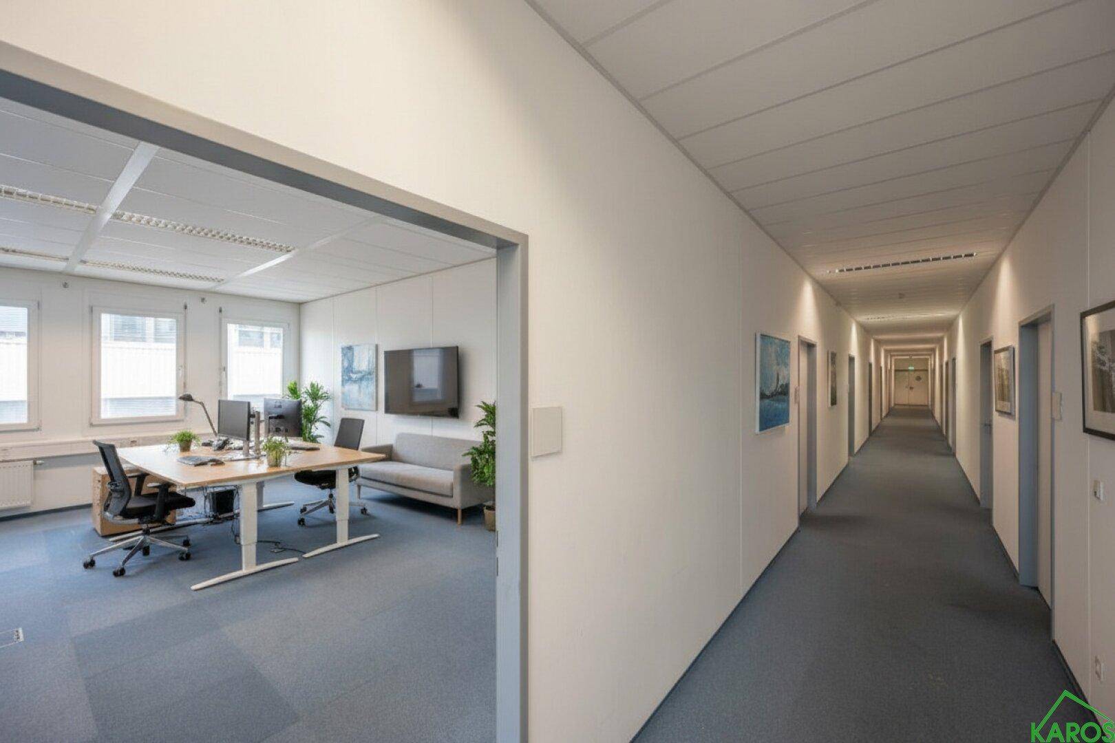 Büroraum mit Arbeitsplätzen und Sofa, der in einen langen, hellen Flur mit vielen Türen führt.