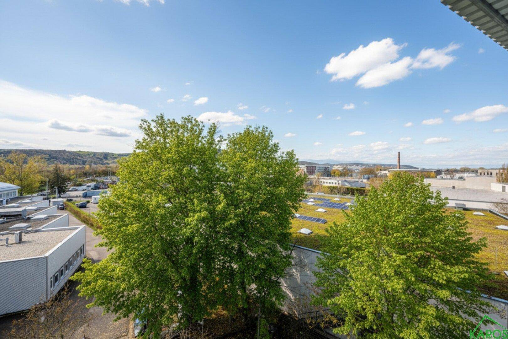 Weitblick über grüne Bäume und Dächer mit Solaranlagen, umgeben von einer industriellen Landschaft unter blauem Himmel.