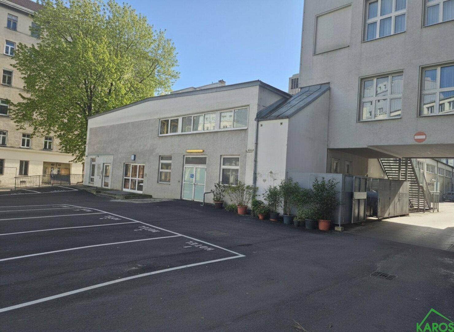 Außenansicht eines modernen Gebäudes mit einem großen Parkplatz davor und angrenzenden Wohngebäuden in einer städtischen Umgebung.