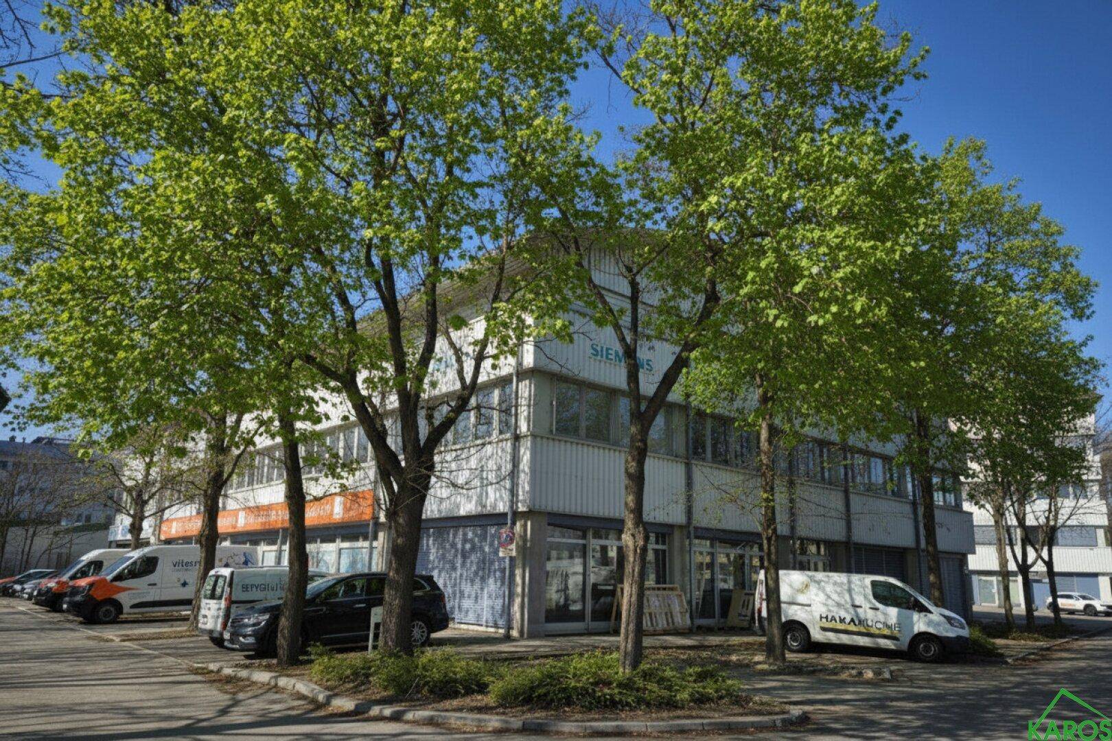 Mehrstöckiges Geschäftsgebäude mit Bäumen im Vordergrund und Parkplätzen auf dem Gelände unter blauem Himmel.