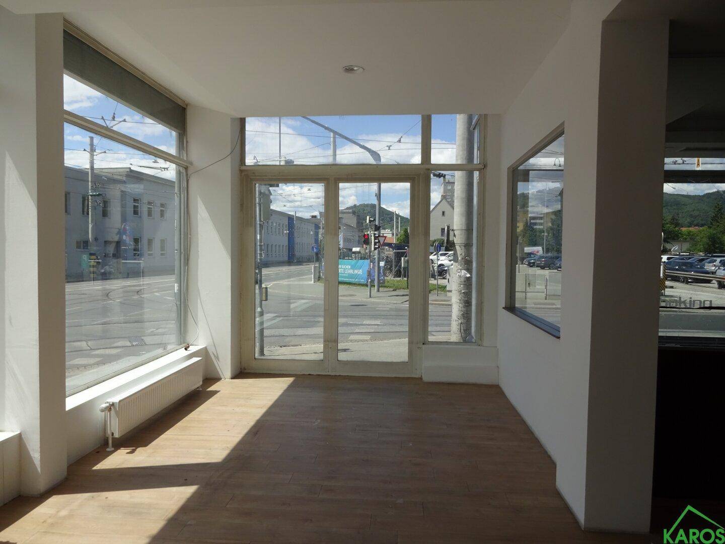 Heller, leerer Geschäftsraum mit großen Fenstern zur Straße und einem gepflegten Laminatboden, der viel Tageslicht hereinlässt.