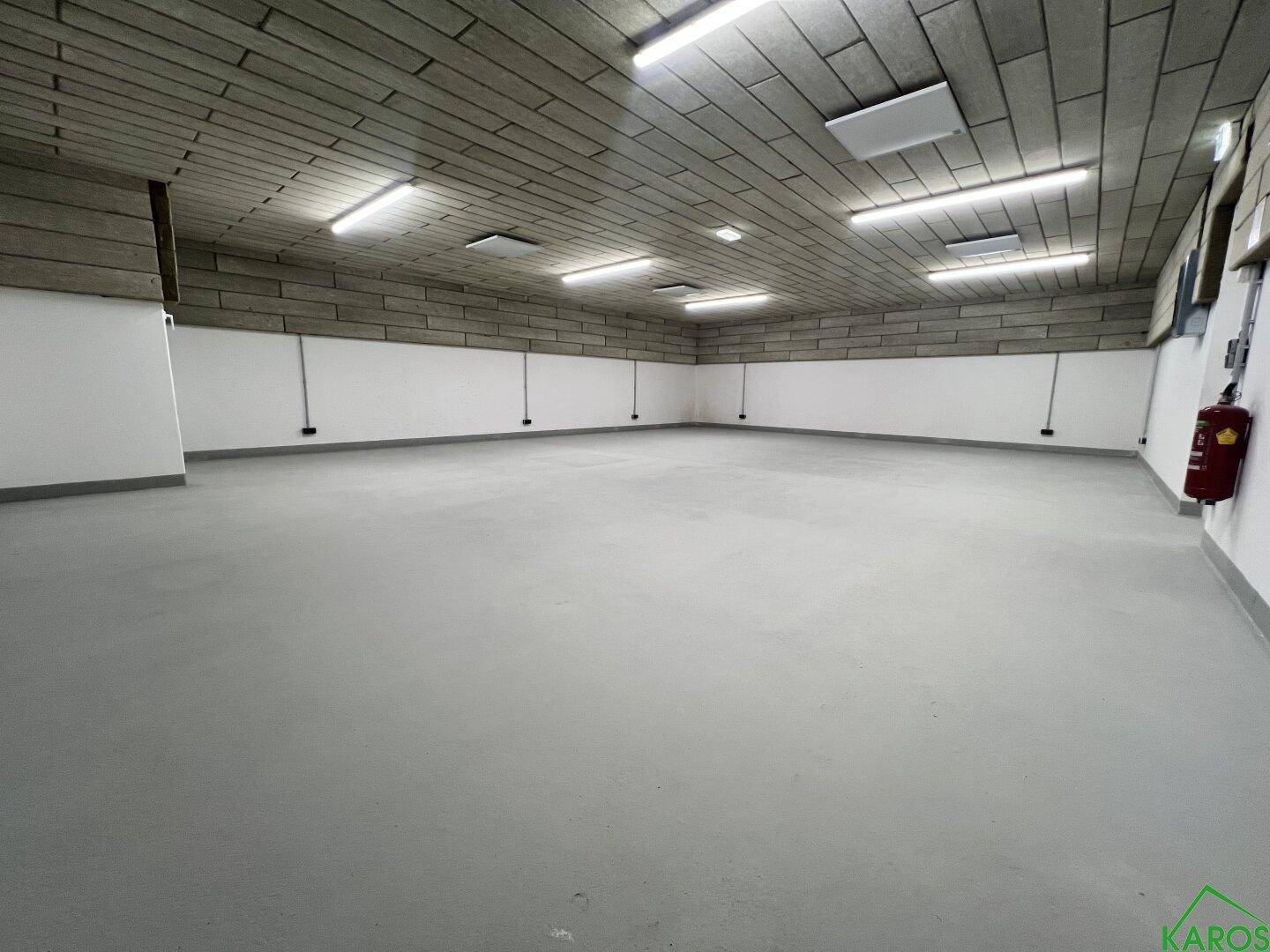 Großer, leerer Raum mit grauem Boden, weißen Wänden und einer Decke mit Akustikplatten und Leuchtstoffröhren.