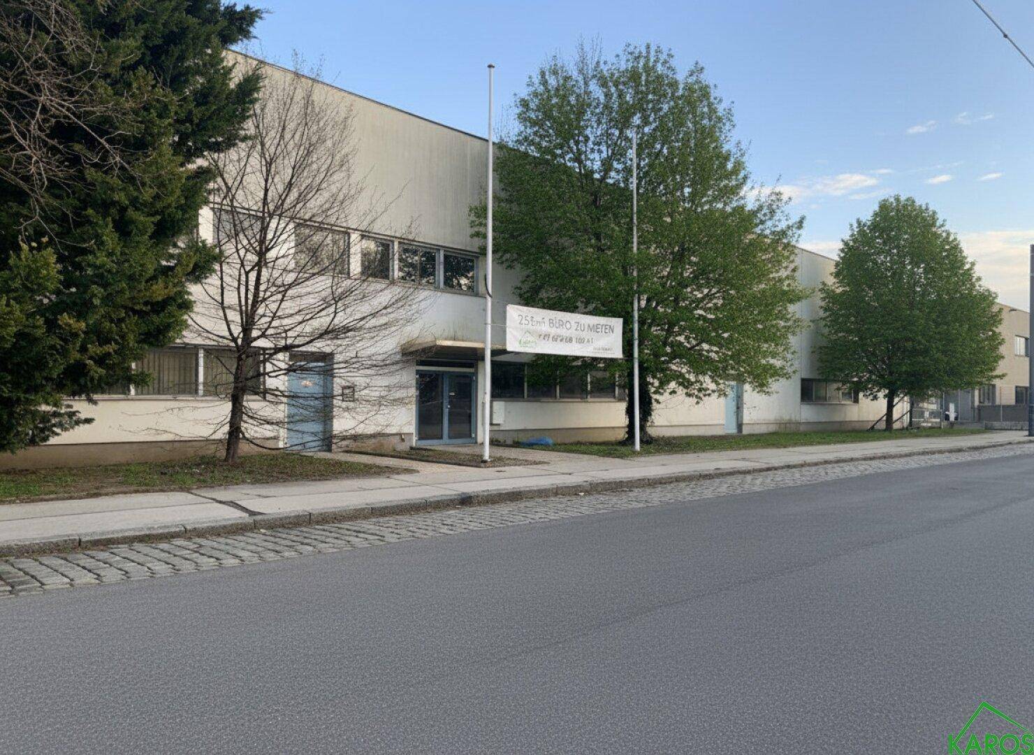 Industrielles Gebäude mit weißer Fassade, vielen Fenstern und Bäumen davor an einem sonnigen Tag.