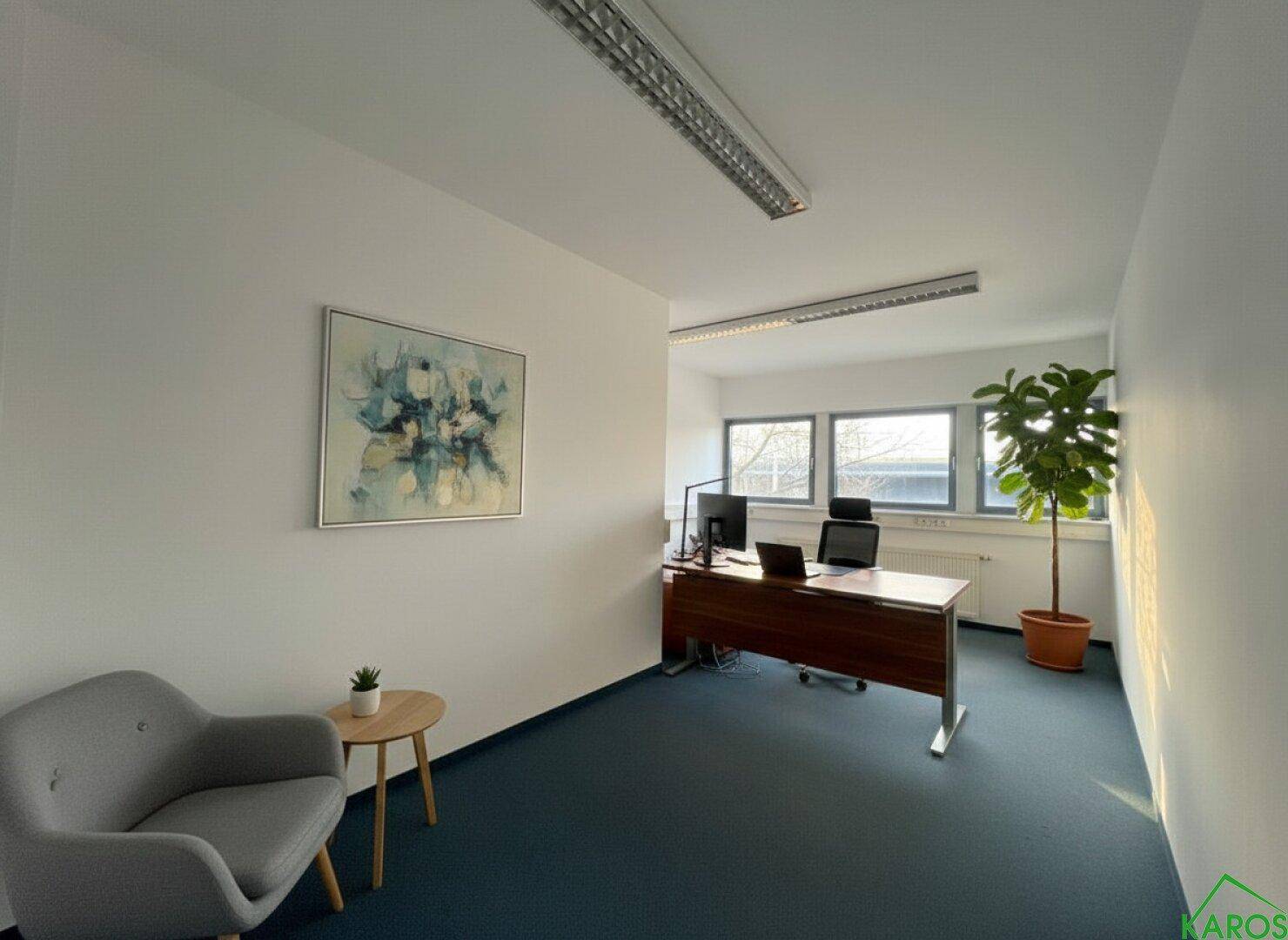 Helles Büro mit blauem Teppichboden, Schreibtisch, Stuhl und einem großen Fenster mit Tageslicht.