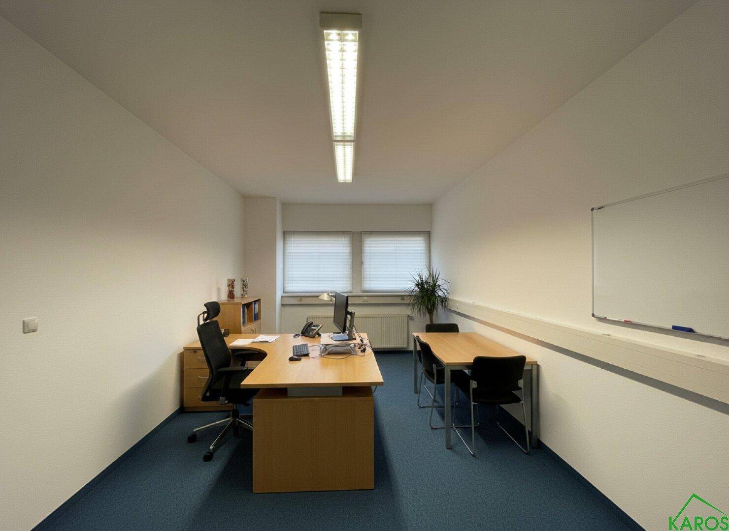 Funktionales Büro mit blauem Teppichboden, Schreibtischen, Bürostühlen und einem Whiteboard an der Wand.