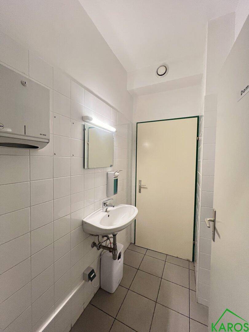 Helle Toilette mit Waschbecken, Spiegel und weißen Fliesen an den Wänden und auf dem Boden.