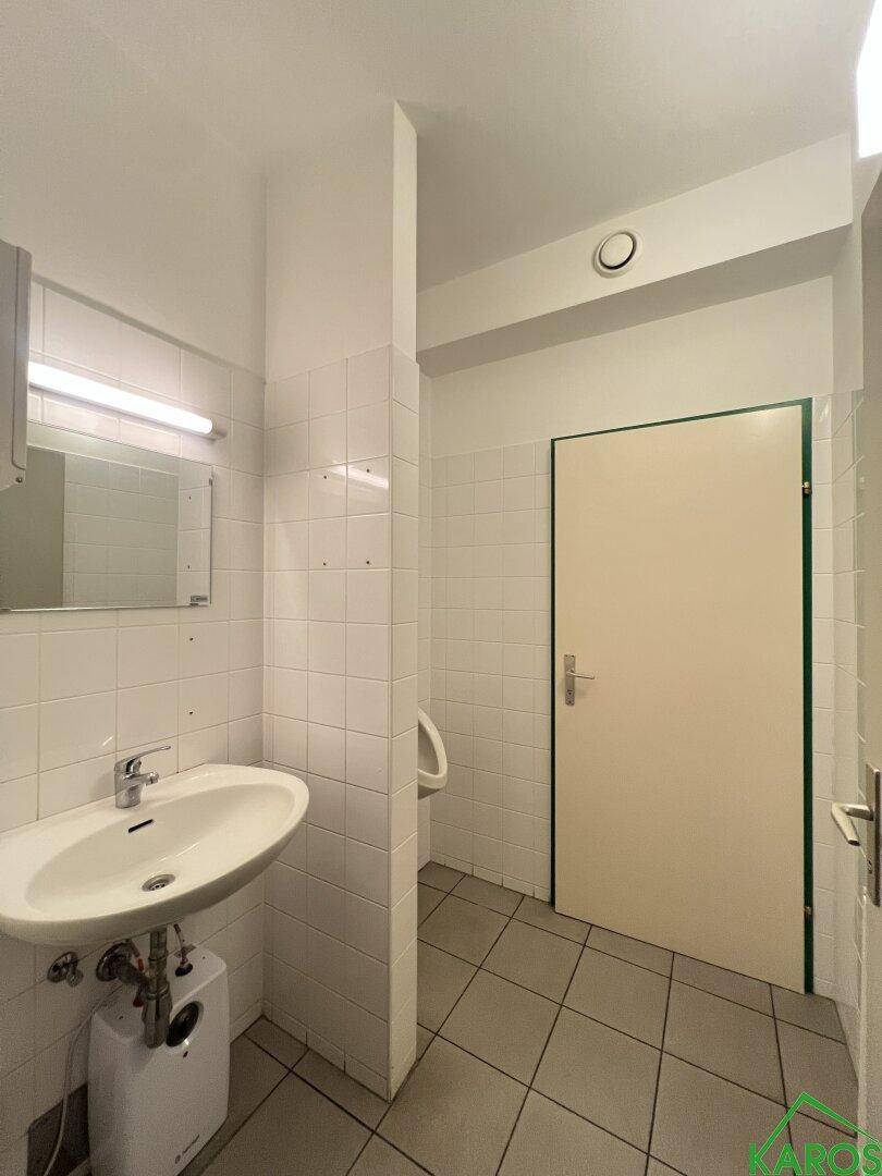 Saubere Toilette mit Waschbecken, Spiegel, weißen Fliesen und einem Urinal im Hintergrund.