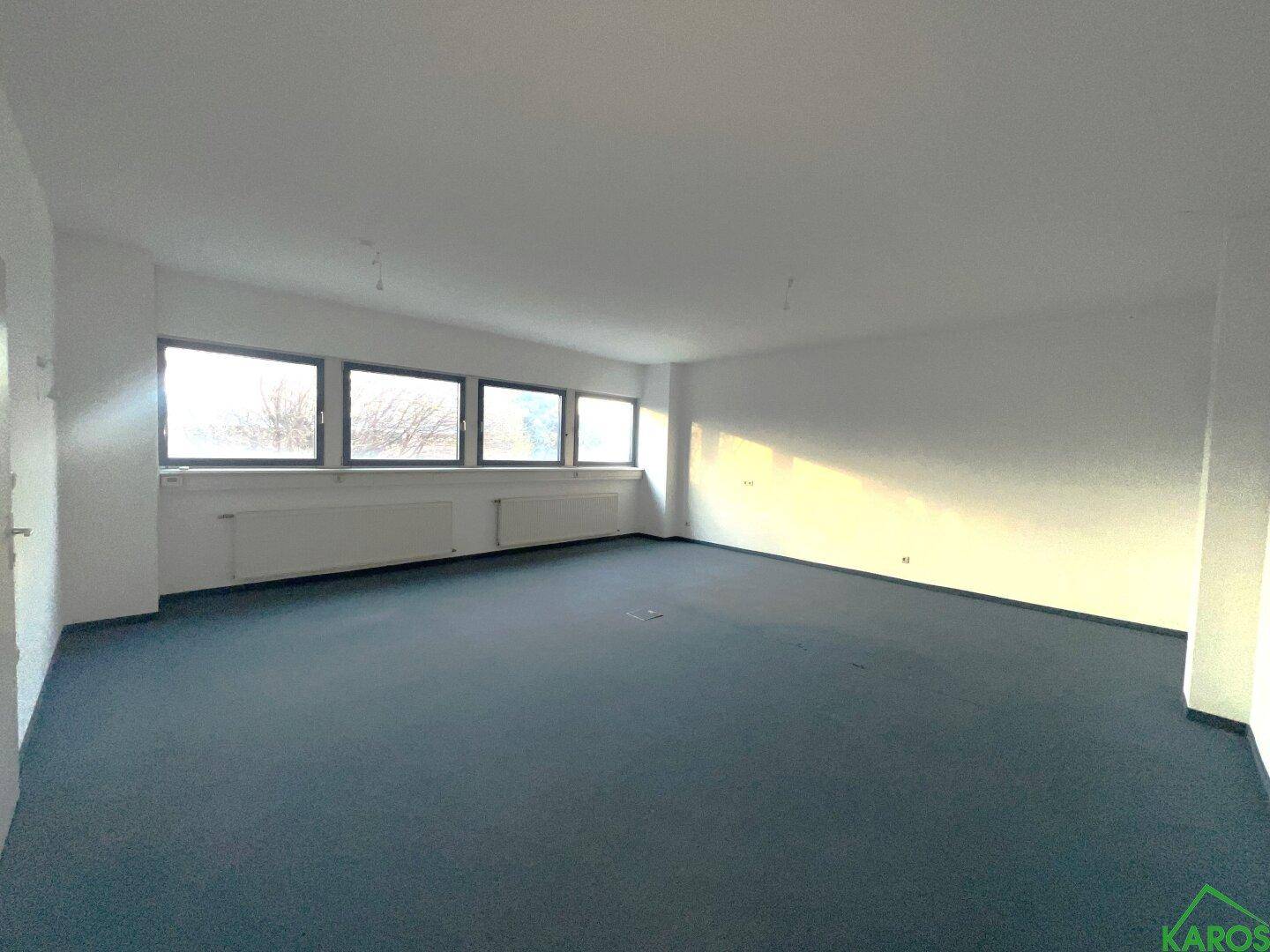 Großer, leerer Büroraum mit blauem Teppichboden, weißen Wänden und einer Fensterfront für viel Licht.