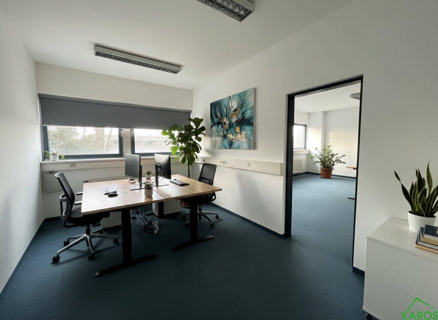 Modernes Büro mit zwei Arbeitsplätzen, blauem Teppichboden und einem großen Fenster mit Rollo.