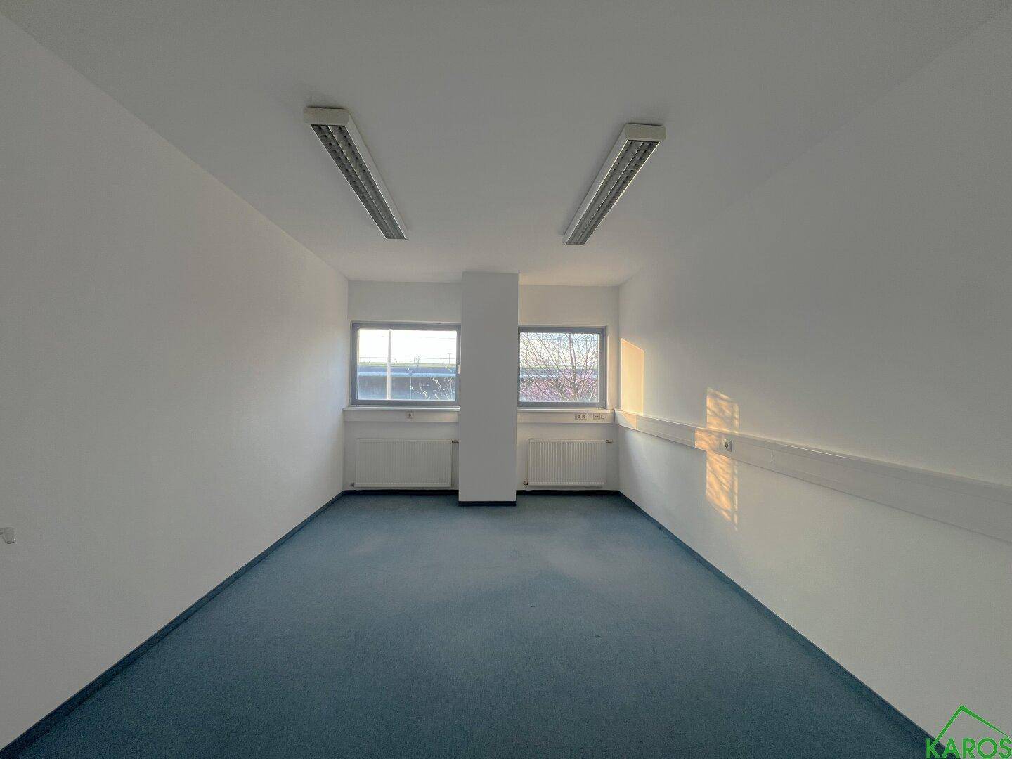 Geräumiger, leerer Büroraum mit blauem Teppichboden, weißen Wänden und Fenstern, die Tageslicht hereinlassen.