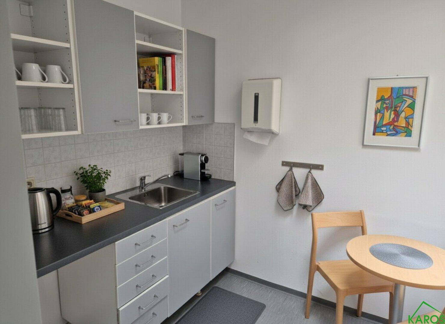 Kleine, funktionale Teeküche mit grauen Schränken, Spüle und einem Essbereich, ideal für kurze Pausen.