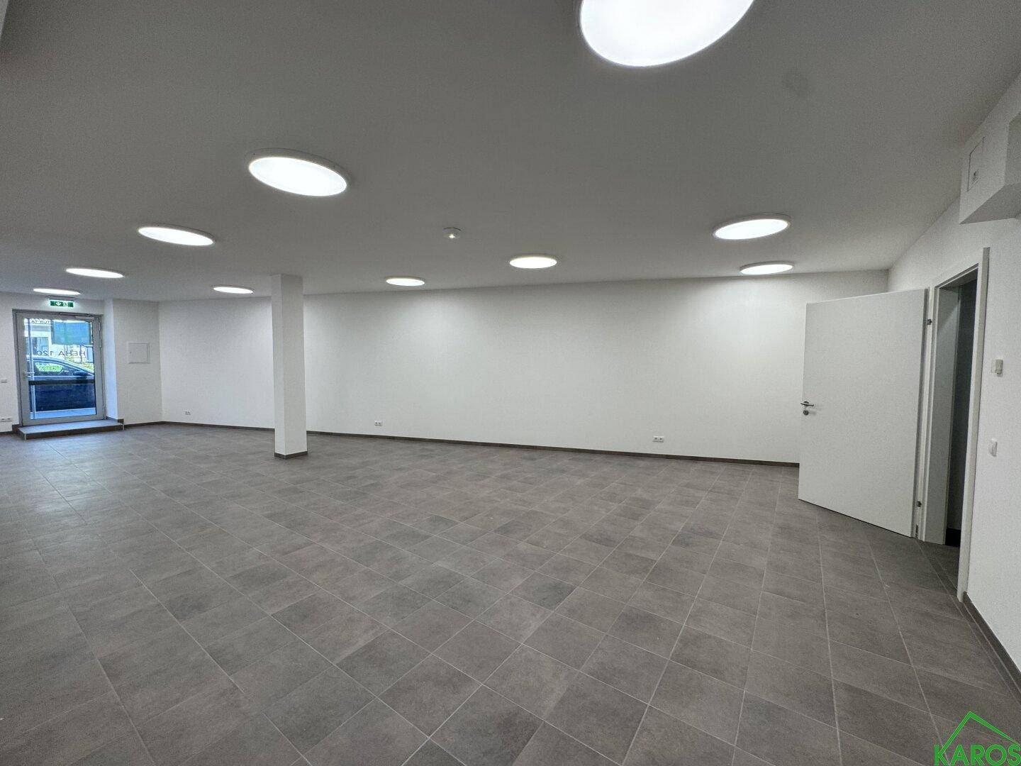 Weitläufiger, heller Büroraum mit grauen Fliesen, weißen Wänden und einer Eingangstür im hinteren Bereich.