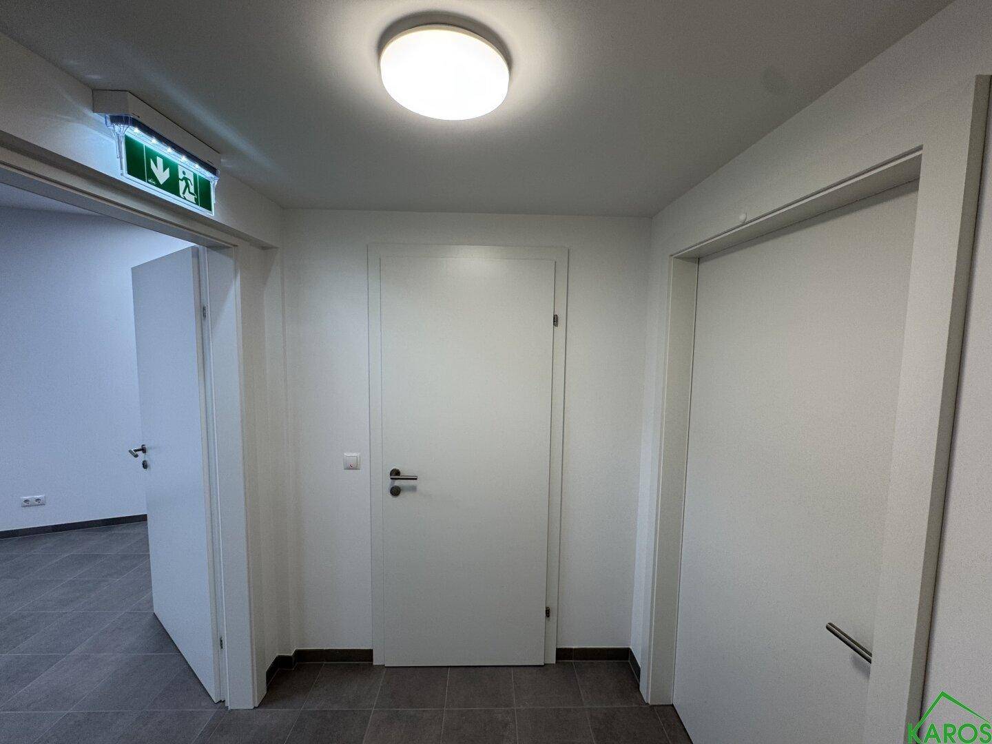 Heller Flur mit mehreren Türen, einem Notausgangsschild und einer runden Deckenleuchte.