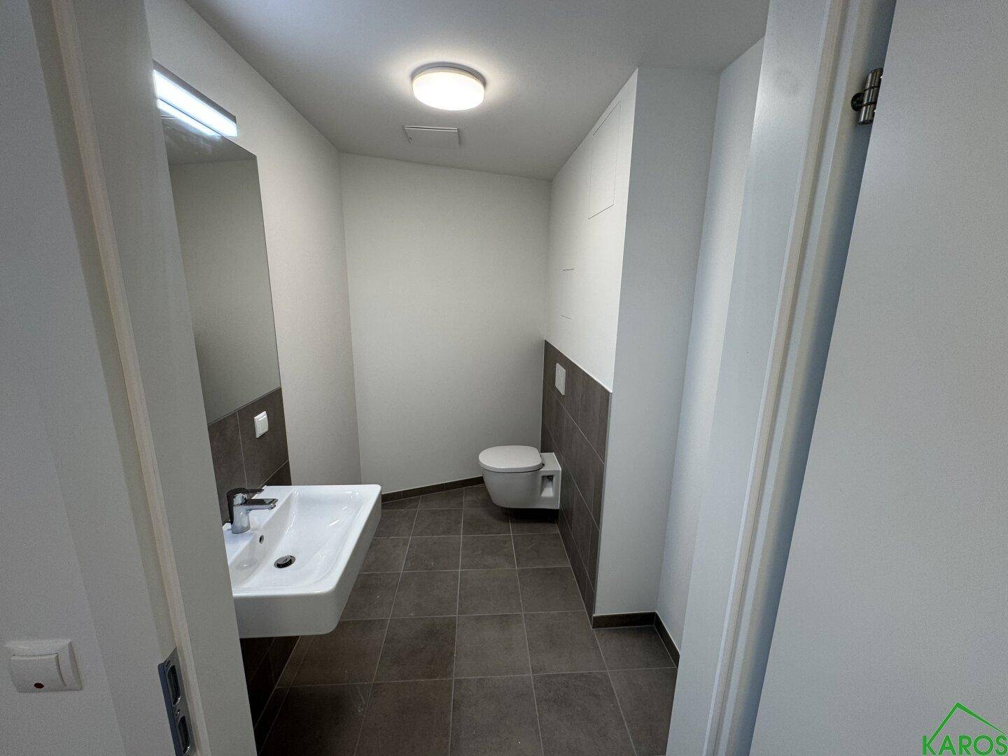 Modernes Badezimmer mit wandhängendem WC, Waschbecken, Spiegel und grauen Fliesen an Boden und Wänden.