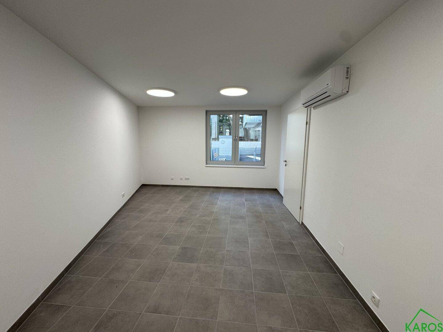 Heller Büroraum mit Fenster, grauen Fliesen, weißen Wänden und einer Klimaanlage, ideal für Einzelbüros.