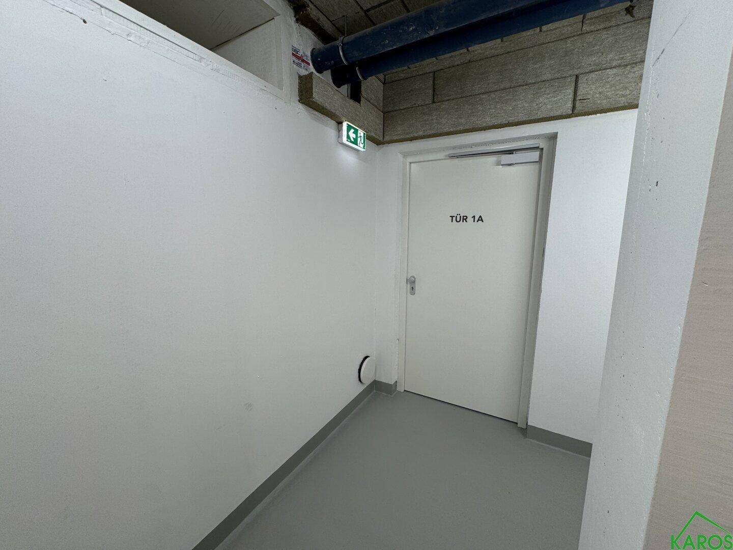 Technikraum oder Kellerbereich mit einer Tür, Notausgangsschild und sichtbaren Rohren an der Decke.