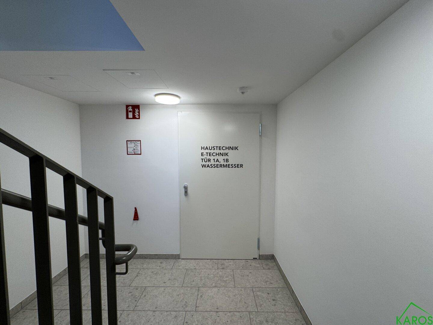 Treppenhausbereich mit einer Tür, die zu den Haustechnikräumen und Wasserzählern führt, in einem modernen Gebäude.