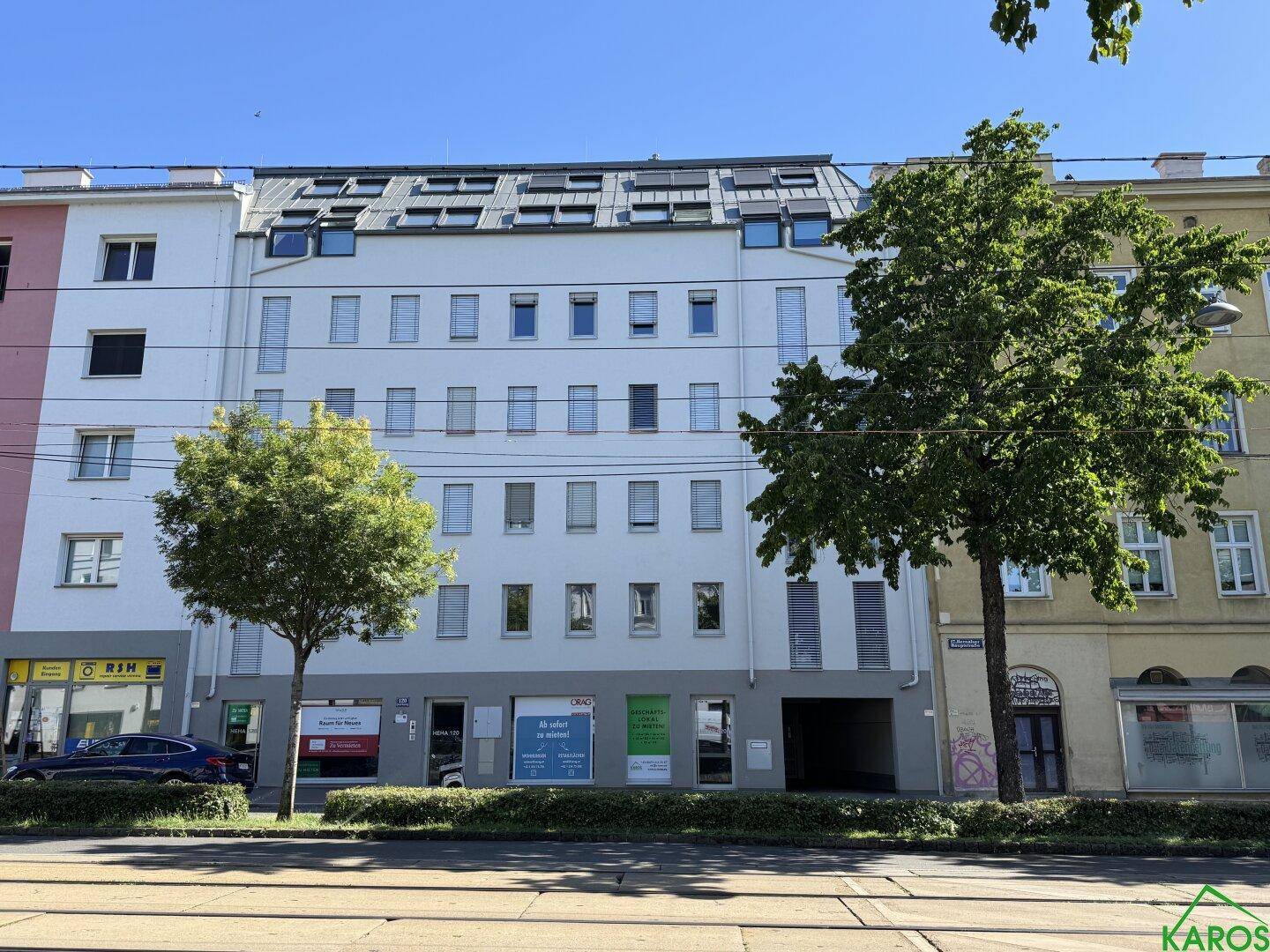 Gesamtansicht der modernen Gebäudefassade mit Geschäften und Wohnungen, umgeben von Bäumen und Stadtleben.