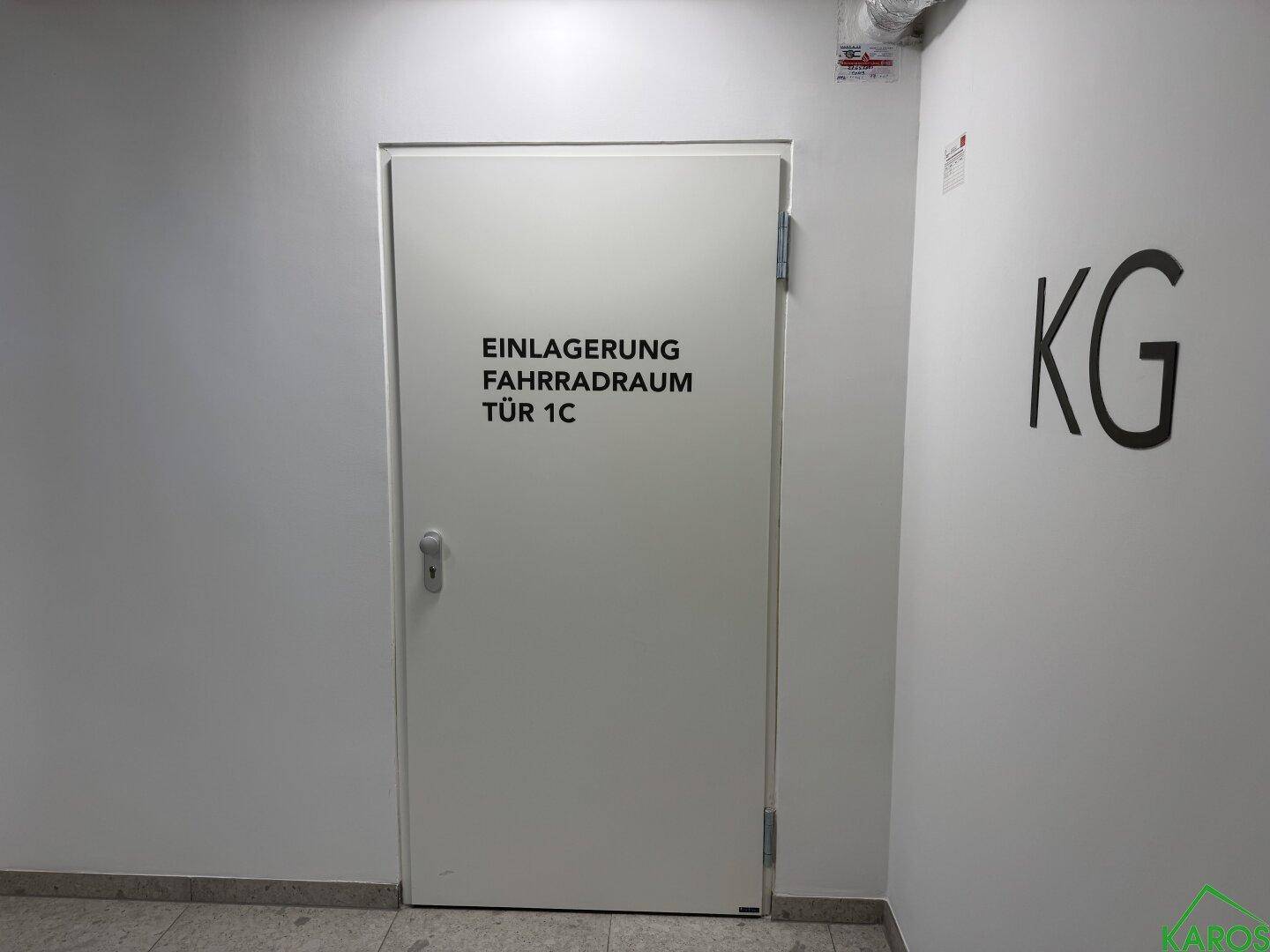 Eine weiße Tür mit der Aufschrift "Einlagerung Fahrradraum Tür 1C" in einem Kellerflur.