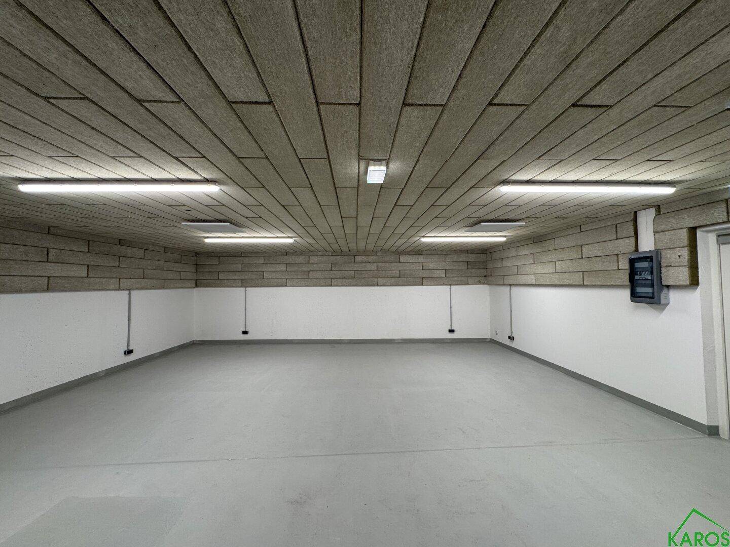 Großer, leerer Lagerraum mit grauem Boden, weißen Wänden und einer strukturierten Decke, mit Elektroverteiler.