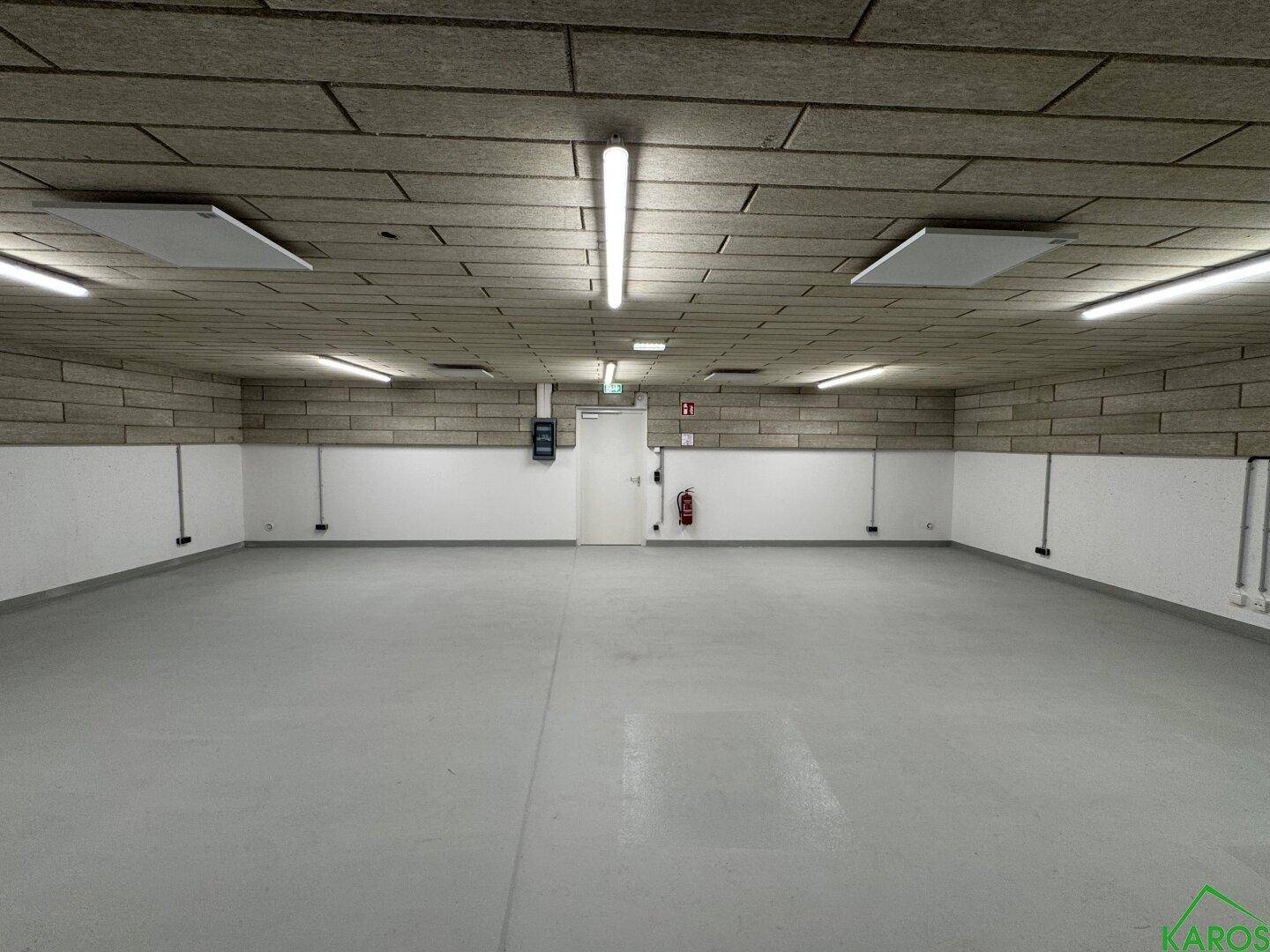 Geräumiger, leerer Lagerraum mit hellem Boden, weißen Wänden und einer strukturierten Decke, mit Tür und Feuerlöscher.
