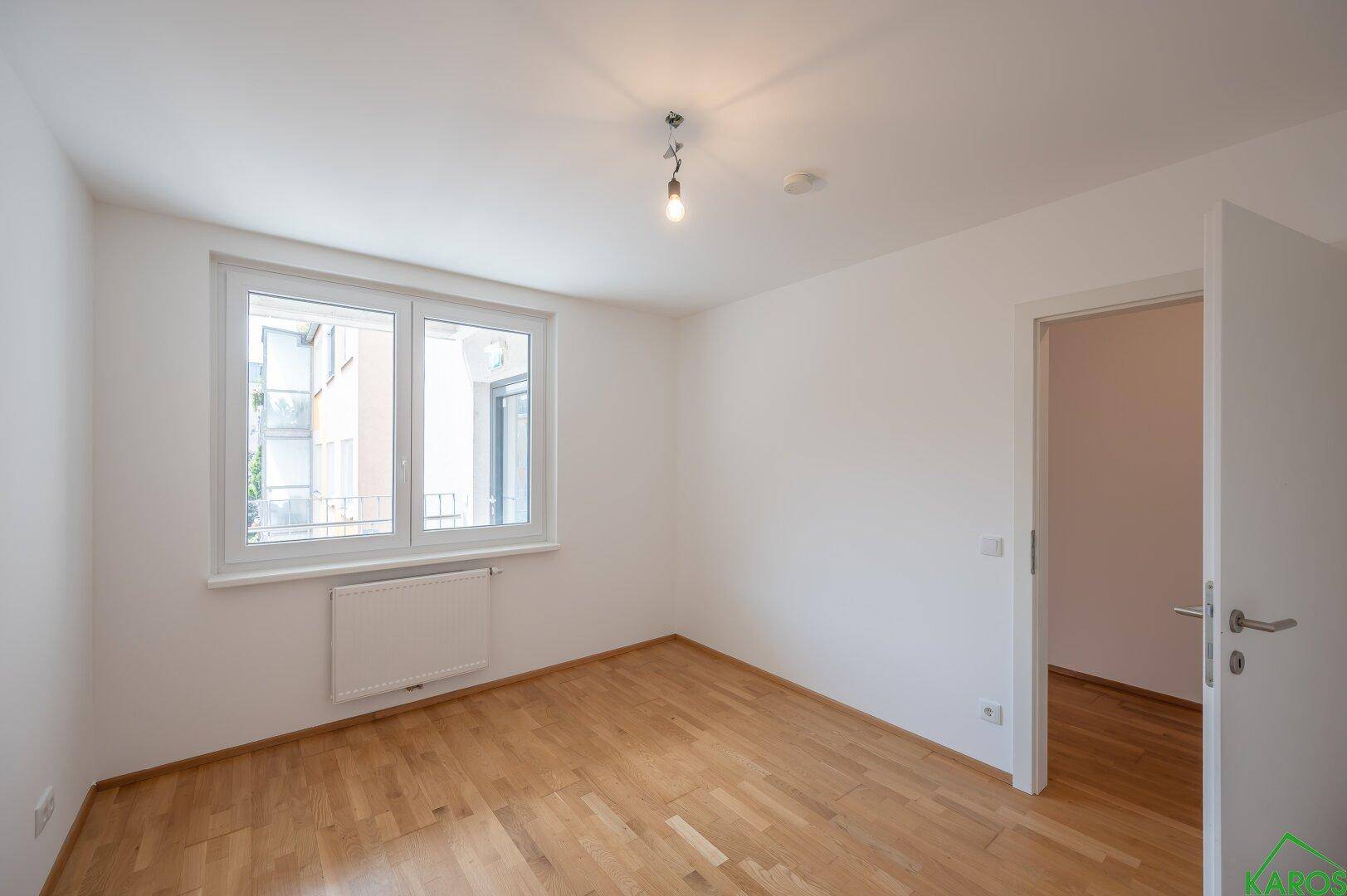 Helles Schlafzimmer mit Holzparkettboden und einem großen Fenster mit Blick nach draußen.