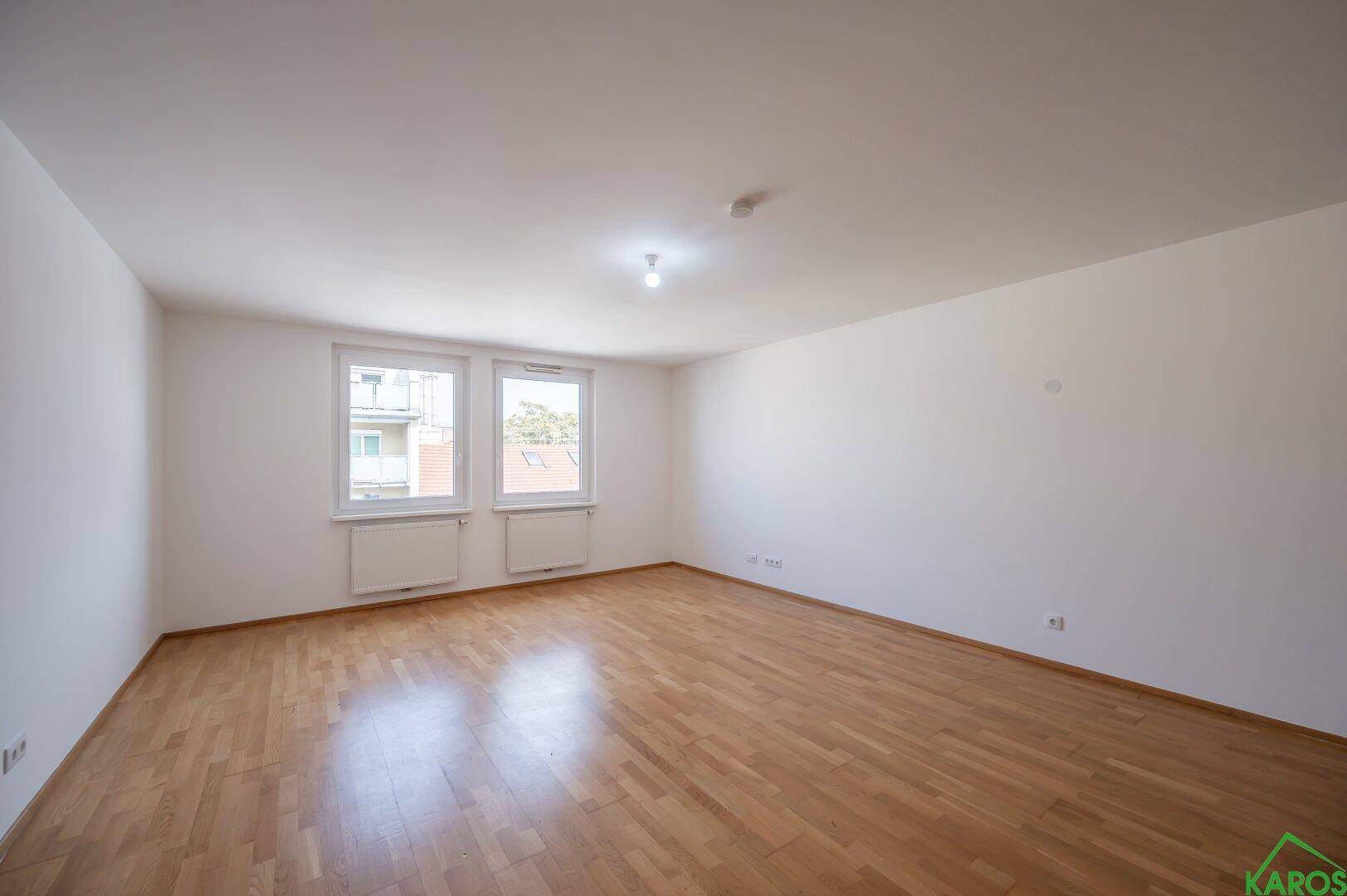 Geräumiges Wohnzimmer mit hellem Holzparkettboden und zwei großen Fenstern.