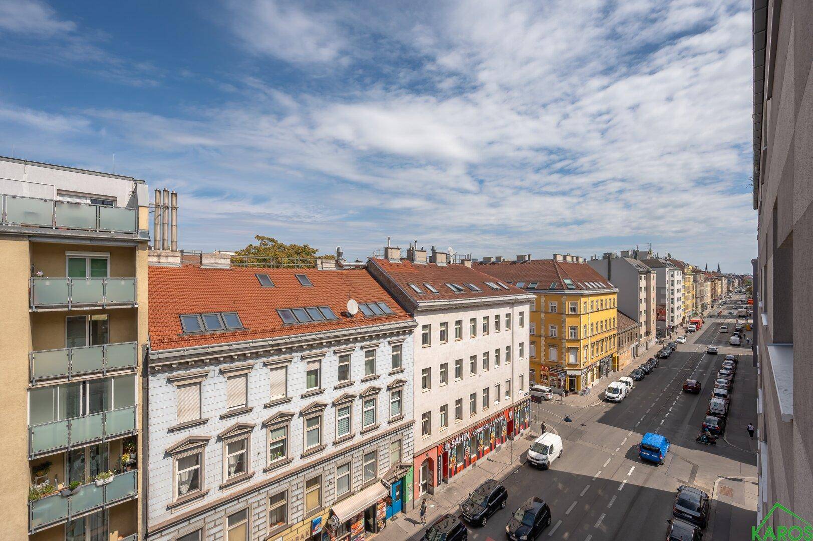 Weitblick über eine belebte Stadtstraße mit Gebäuden und Fahrzeugen unter blauem Himmel.