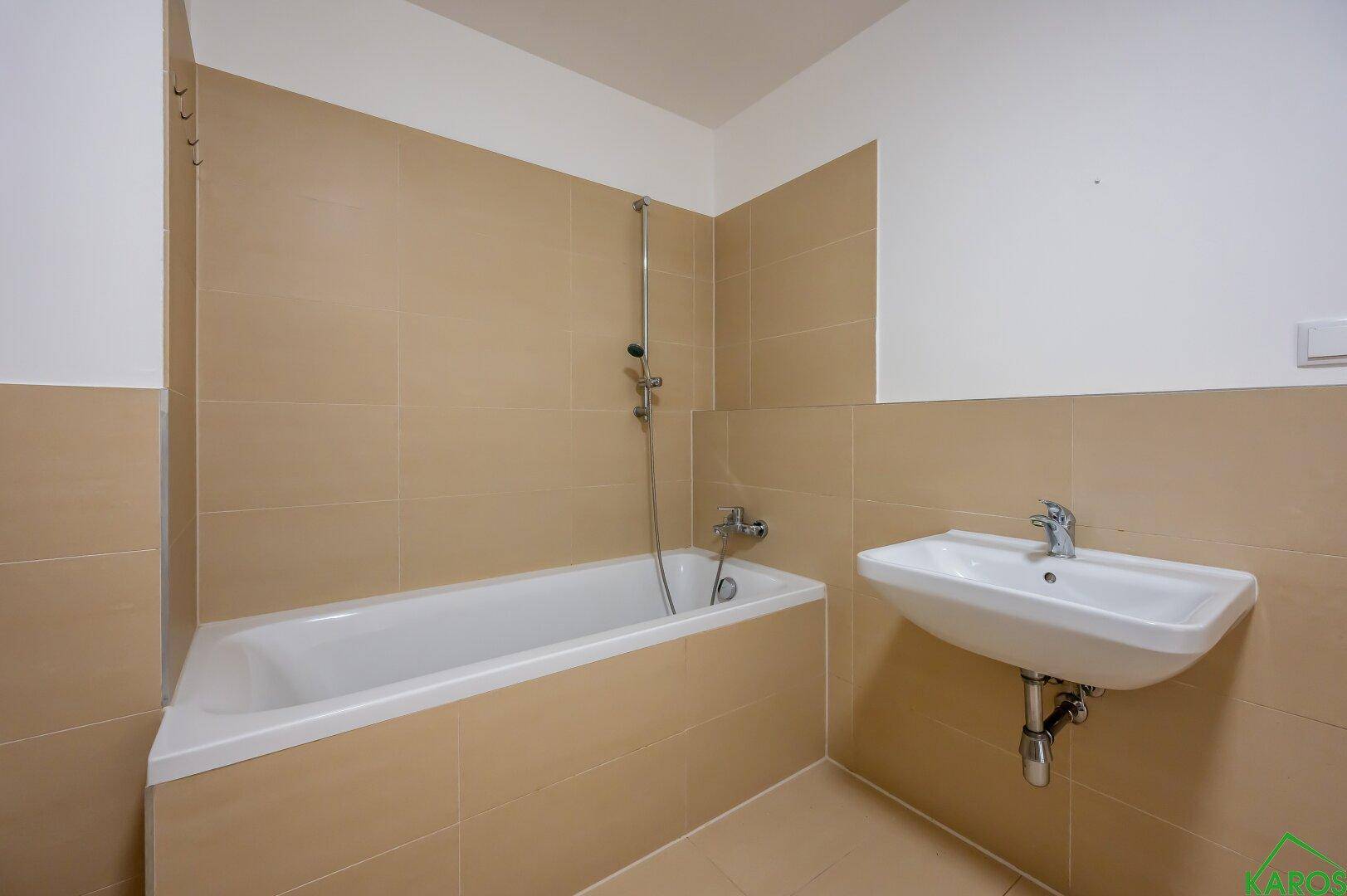 Badezimmer mit Badewanne und Duscharmatur, sowie einem Waschbecken und beigefarbenen Fliesen.