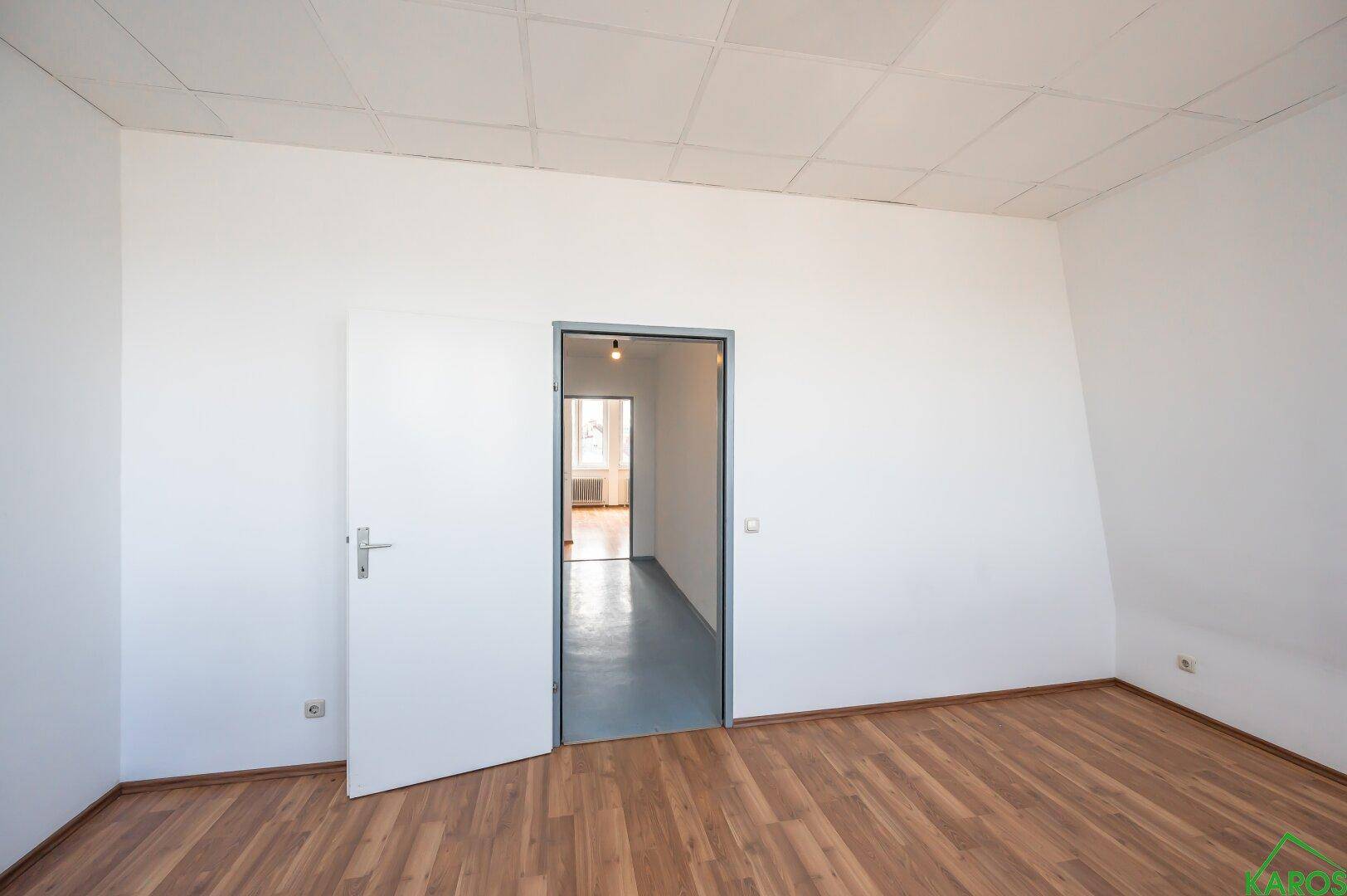 Heller Raum mit Holzfußboden und offener Tür zu einem Flur mit blauem Bodenbelag.