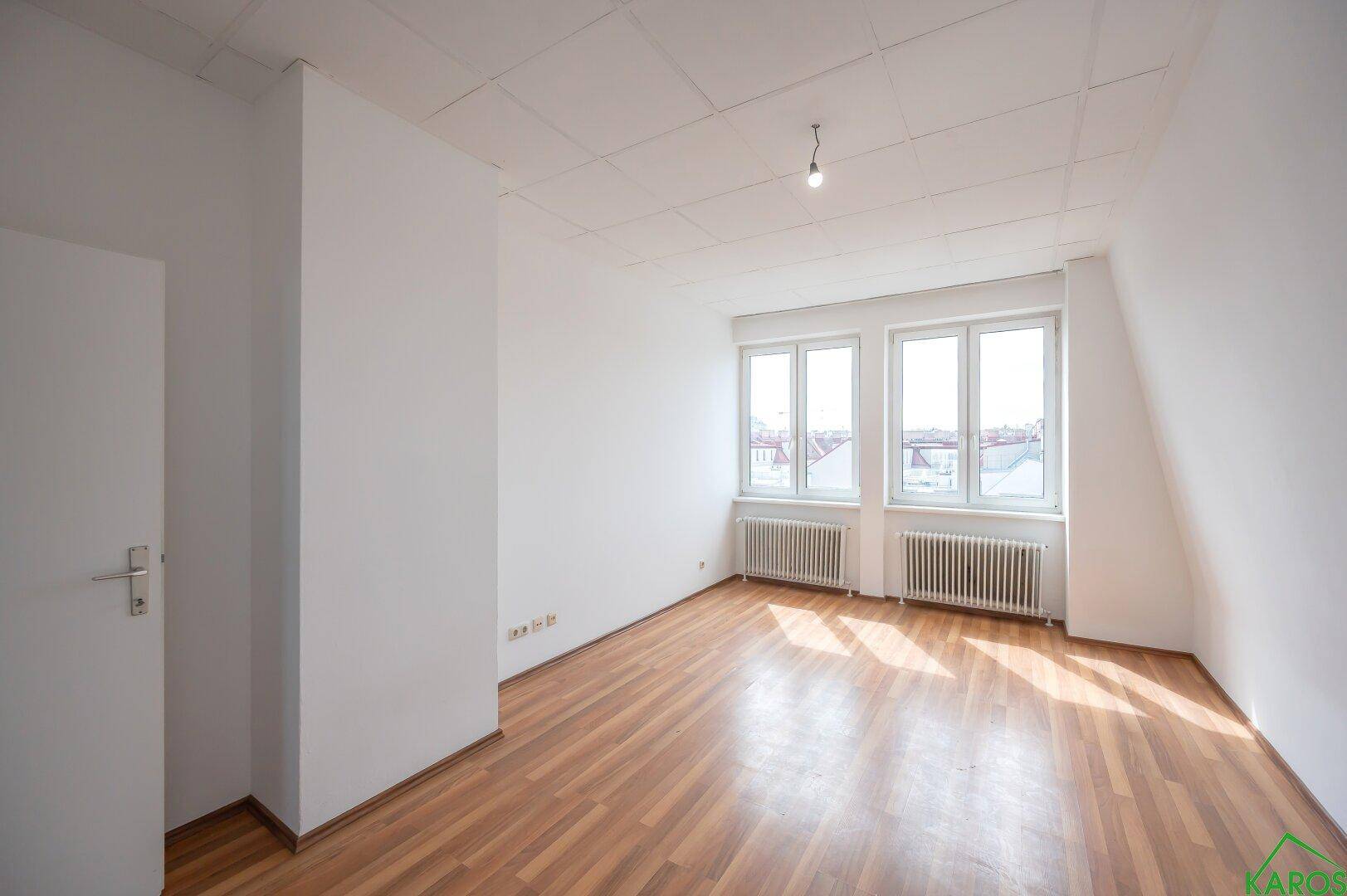 Helles Zimmer mit Holzfußboden und zwei Fenstern, die viel Tageslicht hereinlassen.