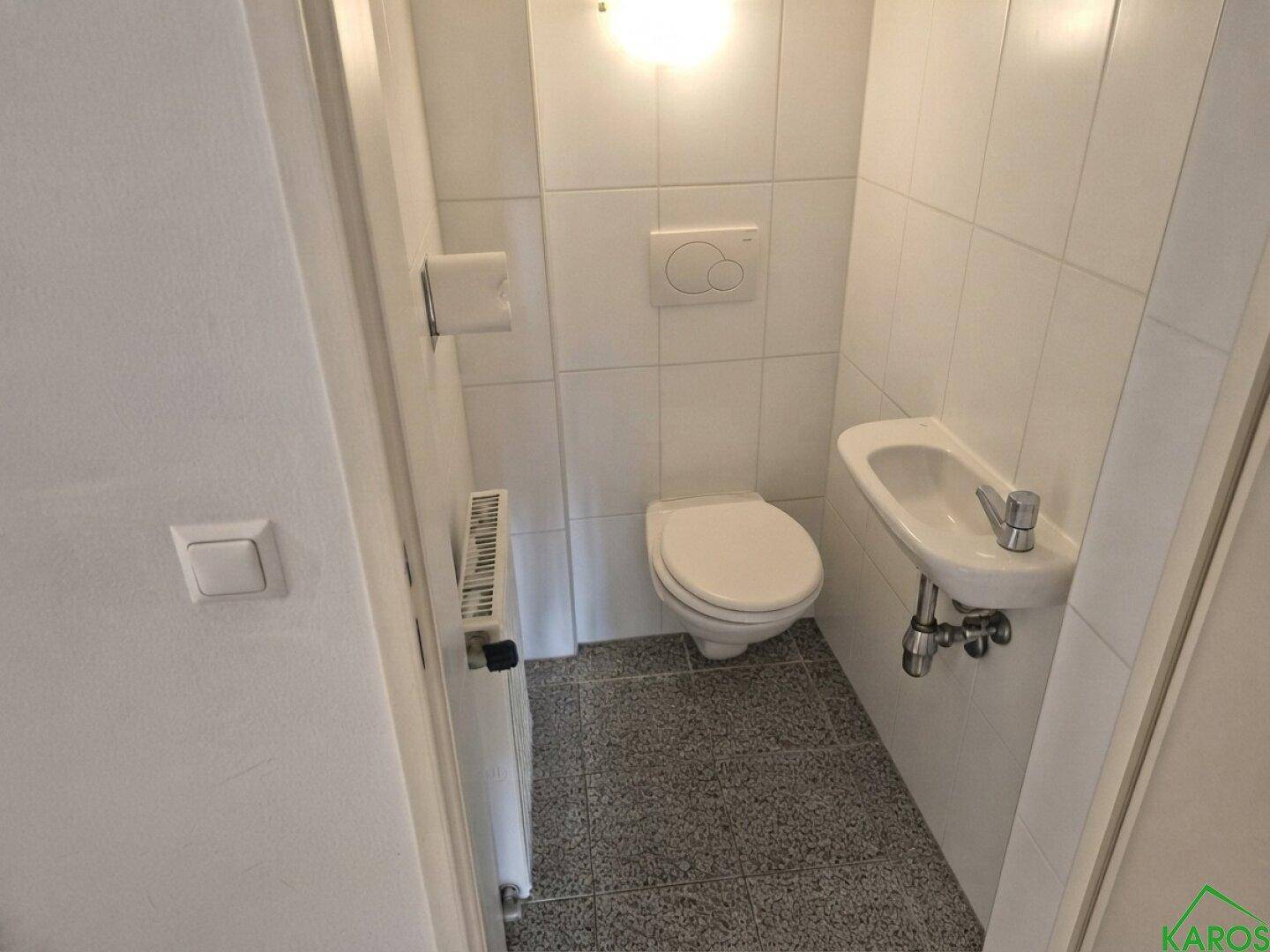 Kleines Gäste-WC mit Toilette, Handwaschbecken und weißen Fliesen an Wänden und Boden.