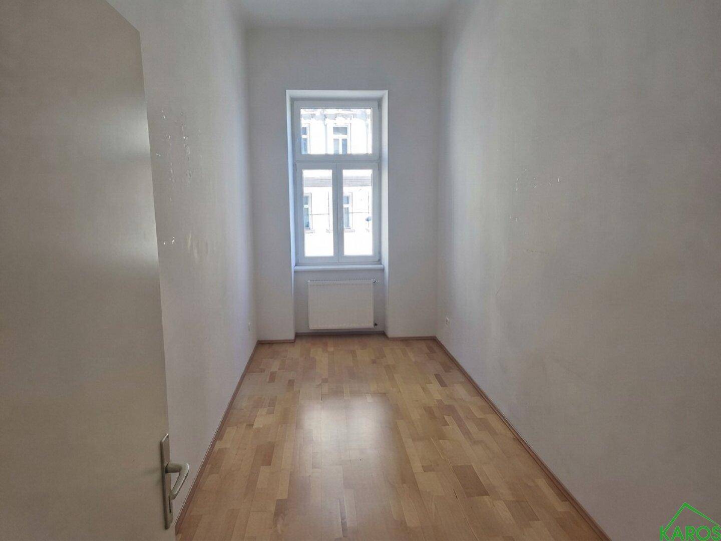Schmales Zimmer mit hellem Parkettboden und einem Fenster, das den Raum erhellt.