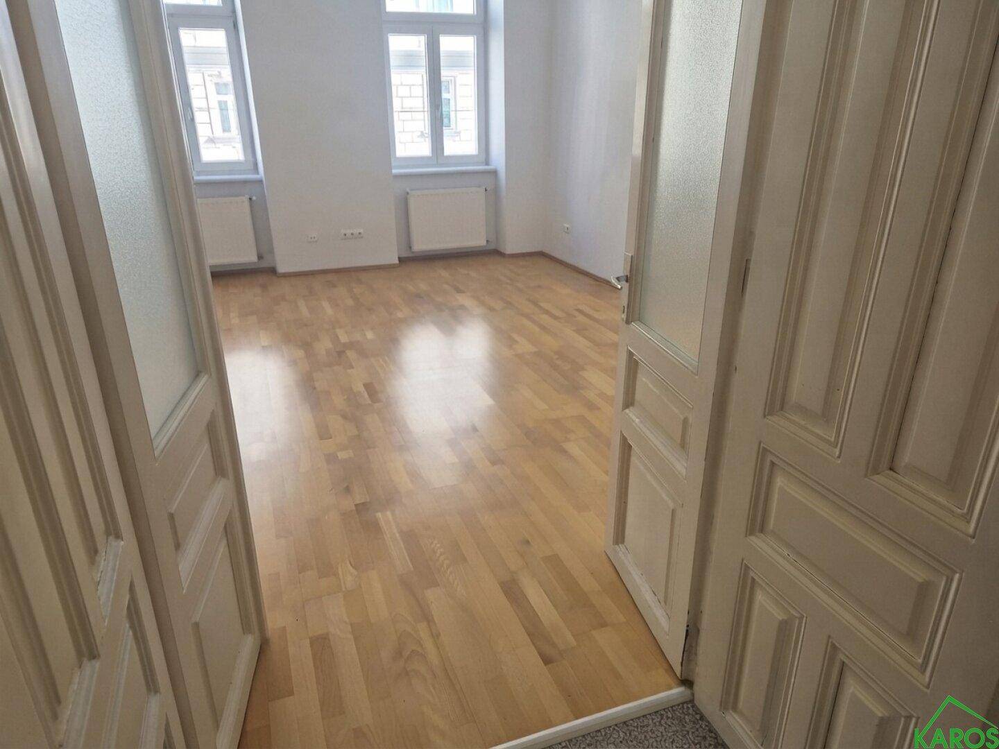 Blick durch eine Tür in ein helles Zimmer mit Parkettboden und zwei großen Fenstern.