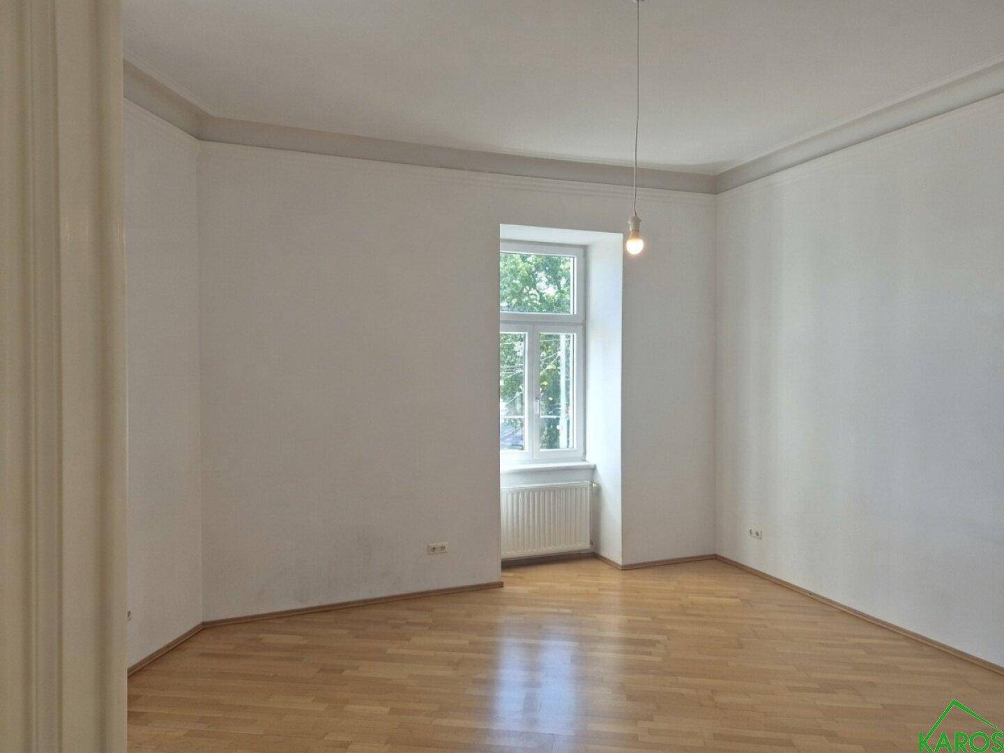 Helles Zimmer mit Parkettboden und einem Fenster mit Blick ins Grüne.