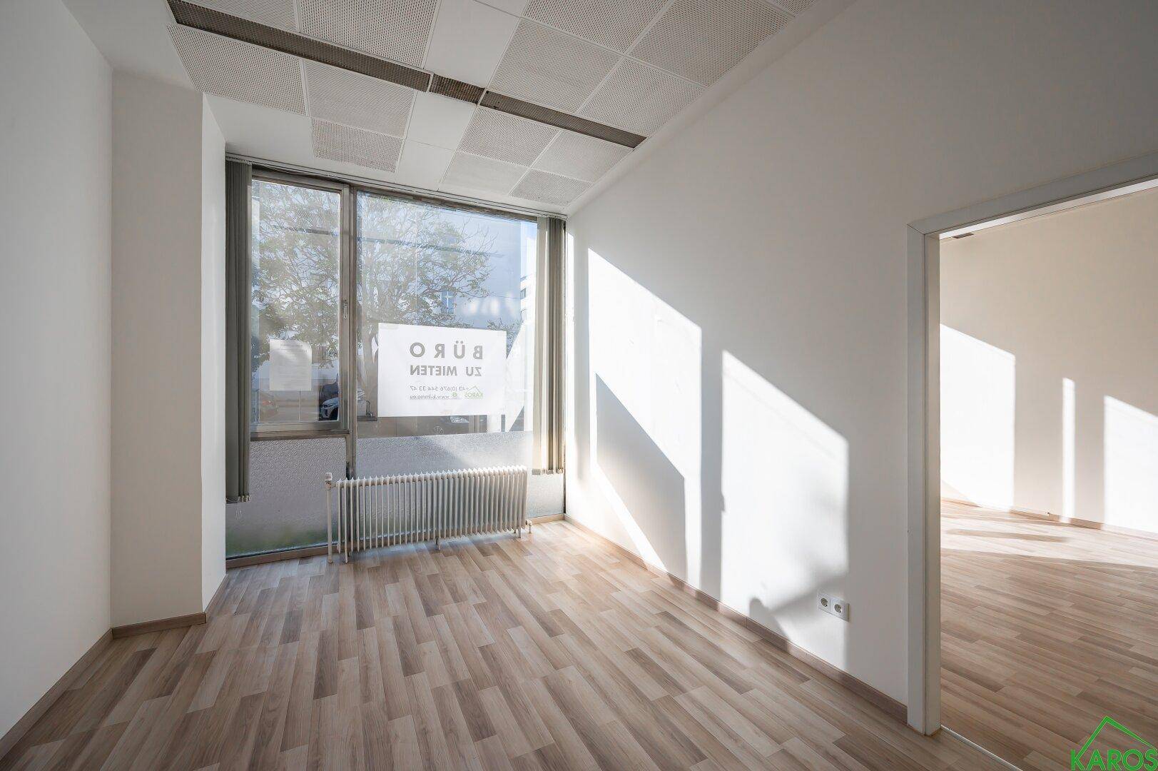Geräumiger Büroraum mit Laminatboden und großen Fenstern, die viel Sonnenlicht hereinlassen.