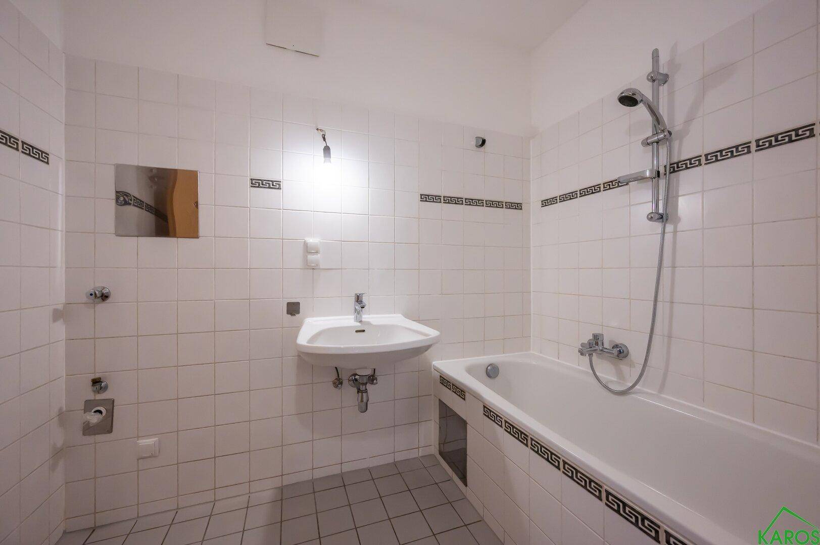 Helles Badezimmer mit weißen Fliesen, Waschbecken und Badewanne mit Duschfunktion.