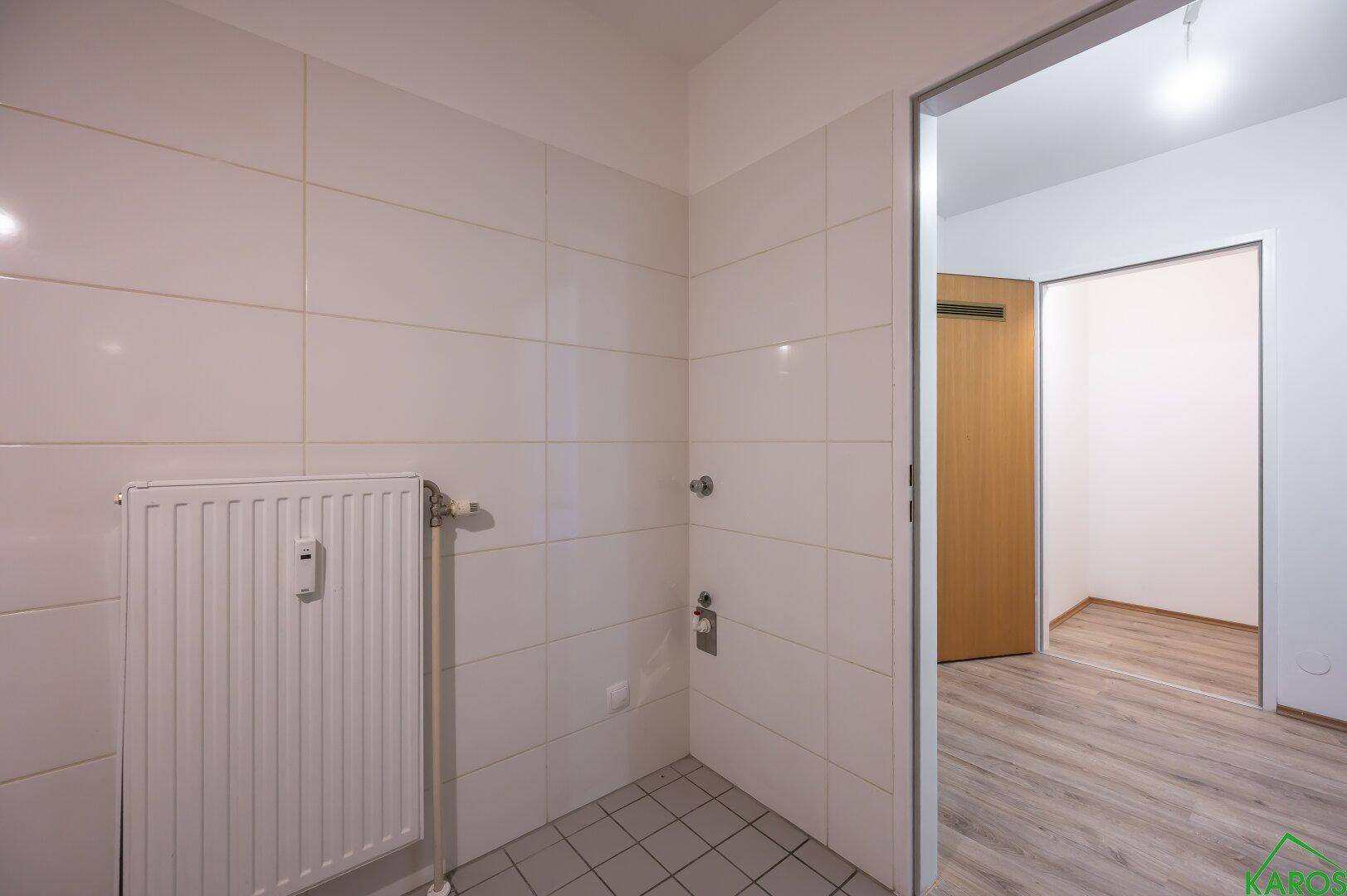 Gefliester Raum mit Heizkörper, möglicherweise ein Hauswirtschaftsraum, mit Zugang zu einem weiteren Zimmer.