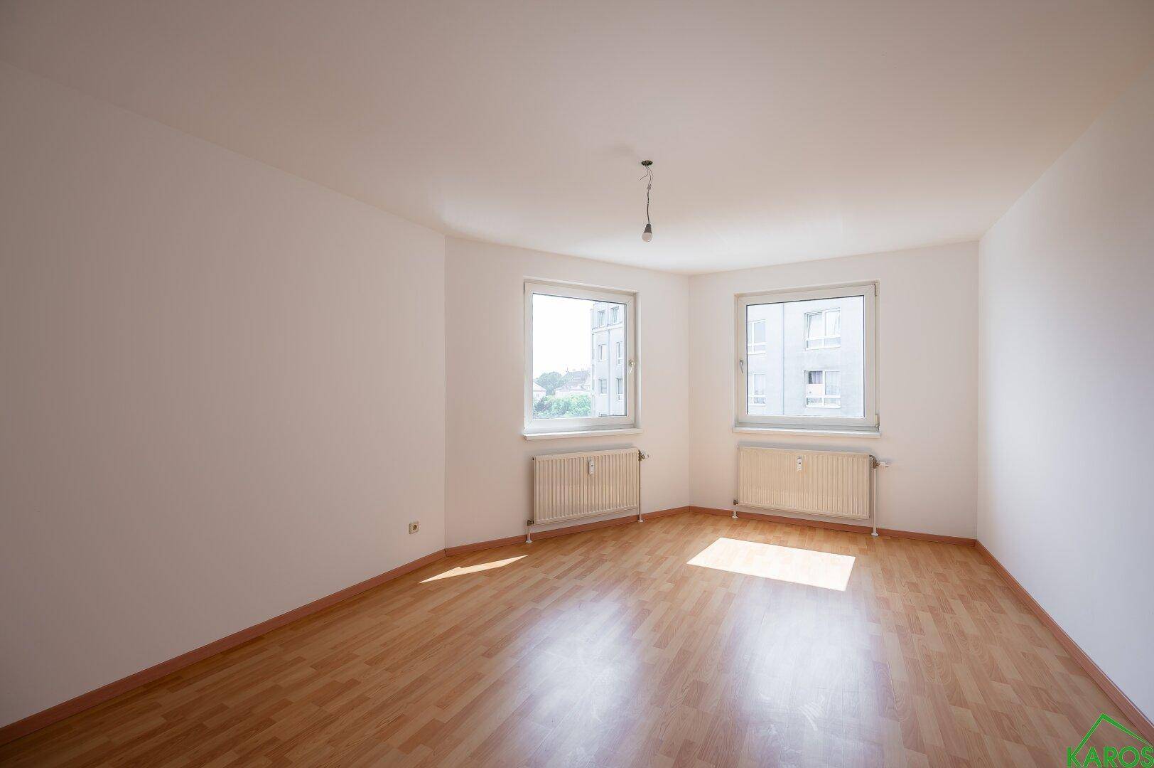 Geräumiges, leeres Wohnzimmer mit Laminatboden und zwei Fenstern, die den Raum hell erscheinen lassen.