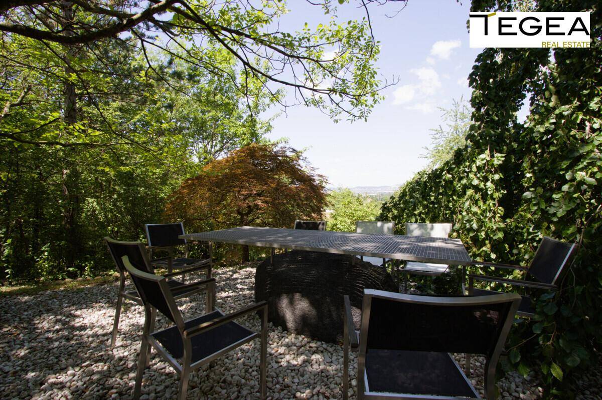 Eine schattige Terrasse mit Esstisch und Stühlen, umgeben von Bäumen und mit Fernblick.