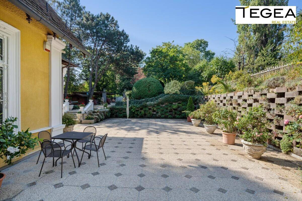 Geräumige gepflasterte Terrasse mit Sitzgelegenheiten, umgeben von Pflanzen und einer Steinmauer.