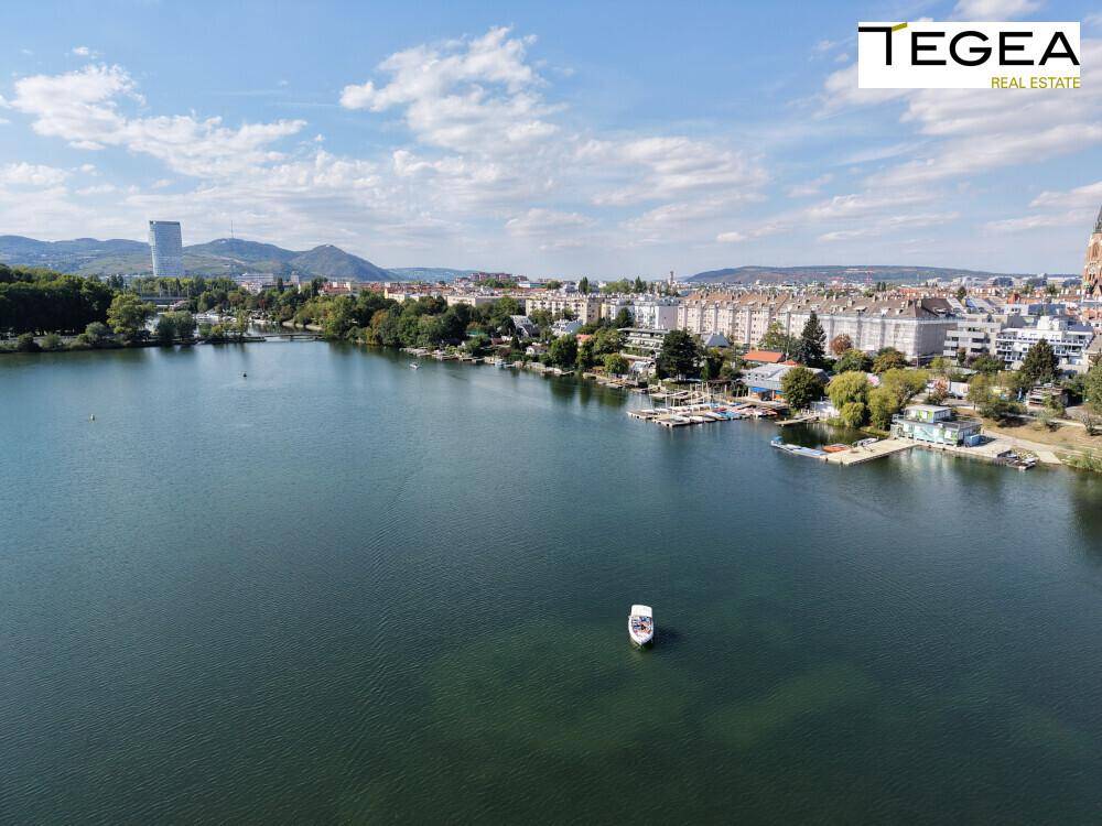 Weitwinkelaufnahme des Sees mit Booten, Uferbebauung und hügeliger Landschaft im Hintergrund.