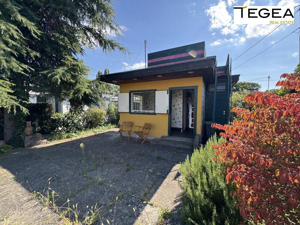 Vorderansicht des gelben Hauses mit kleiner Terrasse und umgebender Vegetation.