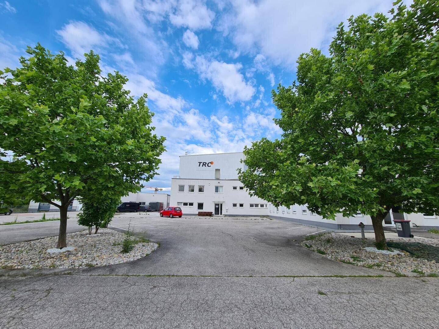 Außenansicht eines modernen Geschäftsgebäudes mit Parkplatz und grünen Bäumen unter blauem Himmel.