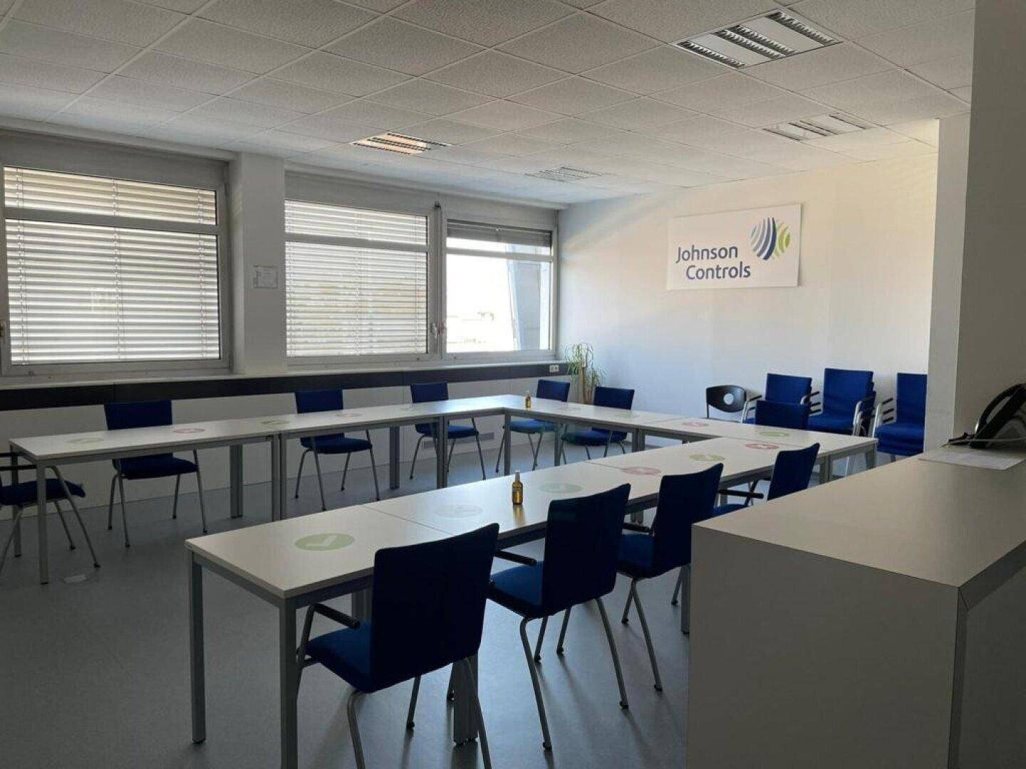 Heller Schulungsraum mit U-förmiger Tischordnung, blauen Stühlen und großen Fenstern.