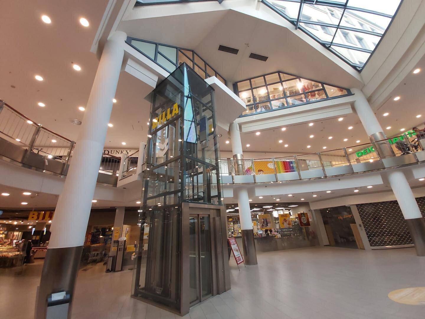 Innenansicht eines Einkaufszentrums mit gläsernem Aufzug, Rolltreppen und einem großen Glasdach.