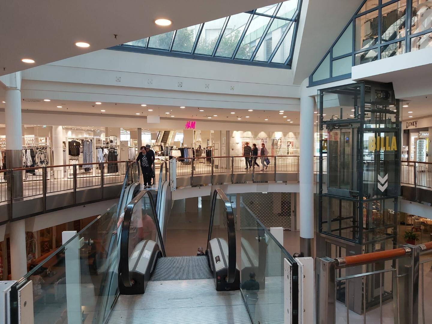 Blick über mehrere Etagen eines Einkaufszentrums mit Rolltreppen, Geschäften und einem gläsernen Aufzug.