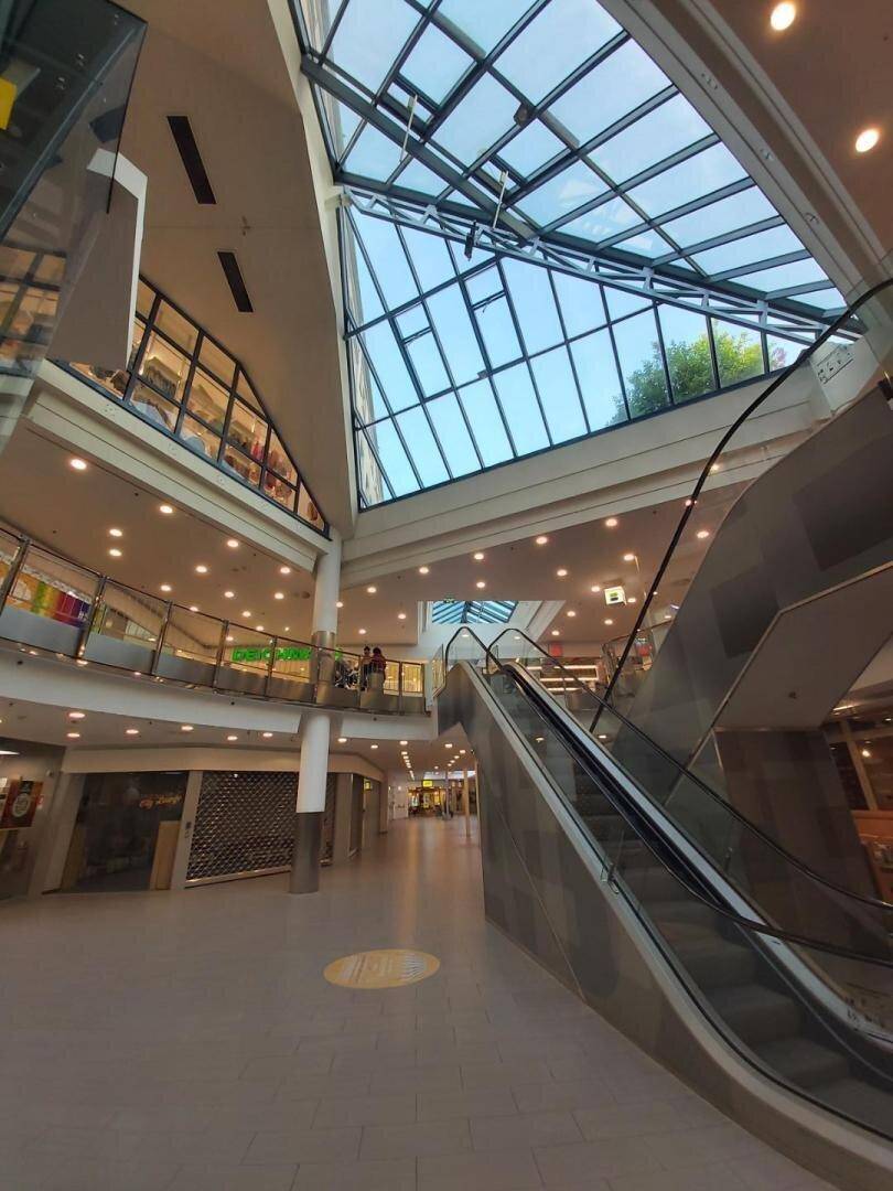 Heller Innenbereich eines Einkaufszentrums mit Rolltreppen und einem beeindruckenden Glasdach, das viel Licht hereinlässt.