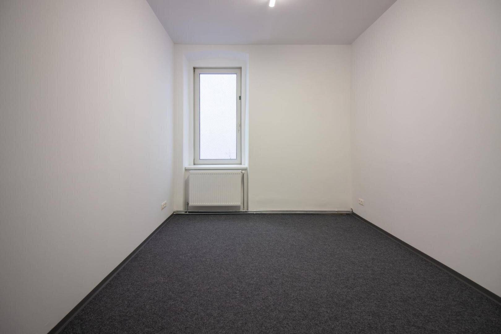 Leerer Büroraum mit weißen Wänden, grauem Teppichboden und einem hohen Fenster mit Heizkörper.