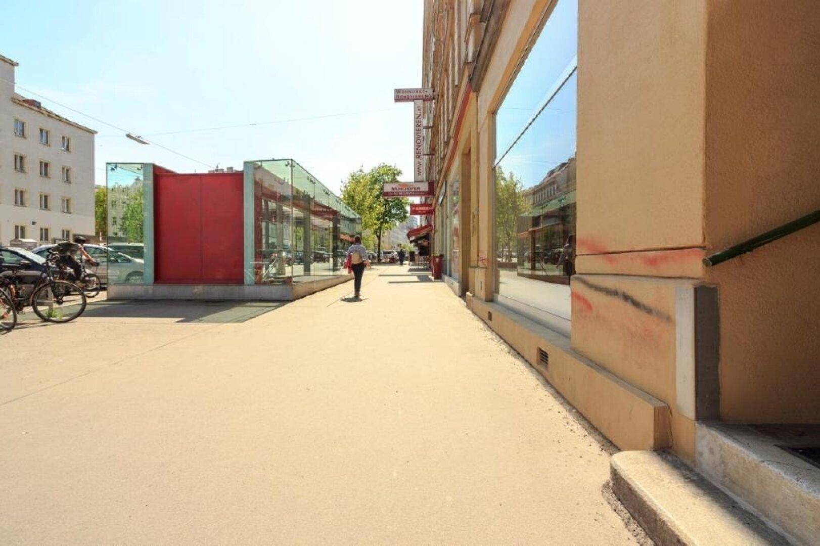 Belebte Straße mit breitem Bürgersteig, Geschäften und einem modernen Glasgebäude, möglicherweise ein U-Bahn-Eingang.